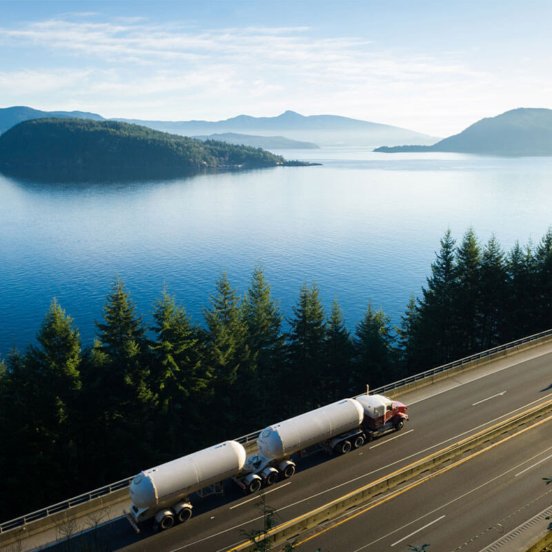 Semi tanker truck driving down a scenic highway with lake, mountains and and sunny sky in the background.